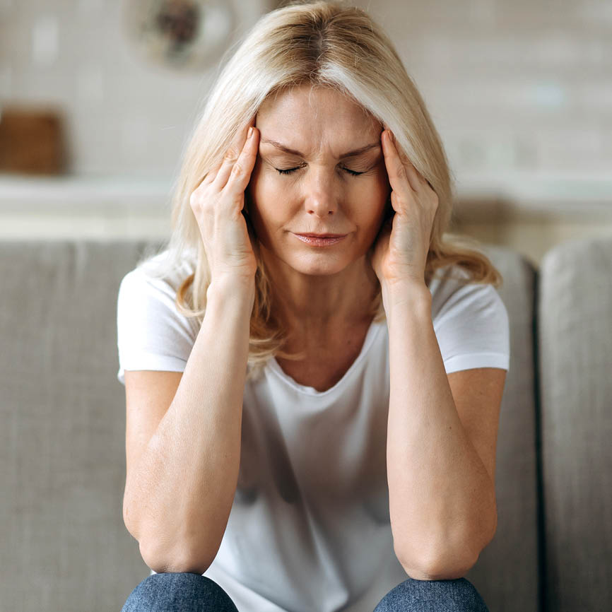 Blonde vrouw in wit T-shirt en spijkerbroek houdt haar handen tegen haar hoofd, wat hoofdpijn klachten suggereert.